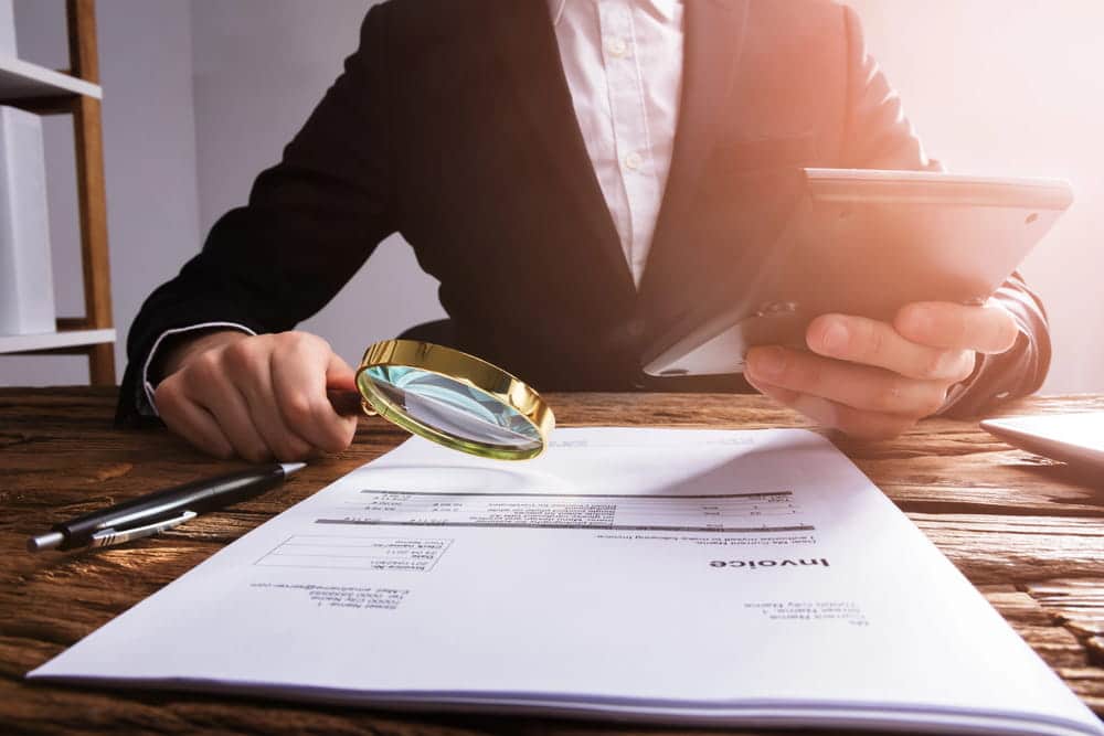Image is of a professional analyzing documents with a magnifying glass and tablet, showing careful document review processes used in asset investigations