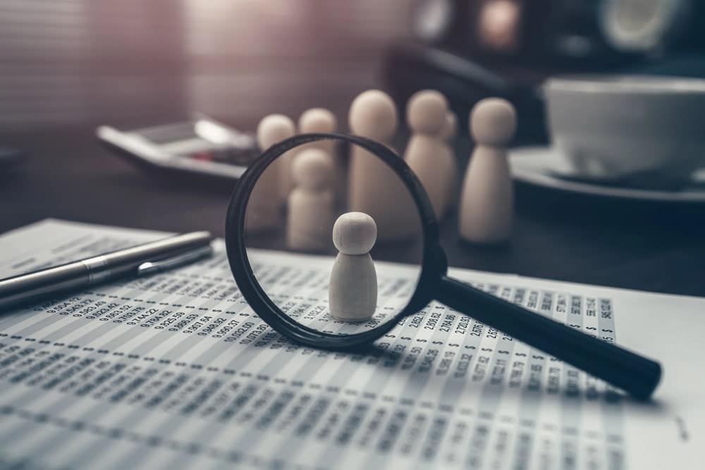 Image is of a magnifying glass highlighting an individual figure on financial records, concept of asset search used by attorneys to identify ownership and recoverable assets.